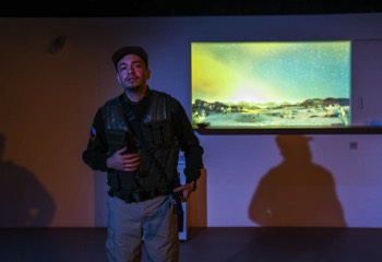 A Fight Against Production Image. A border guard, Adrián (Sebastian Orozco) stands alone in full military gear. He has an automatic weapon hanging over his shoulder. He looks out in the moonlight. There's a projection of a desert scape through a window behind him.
