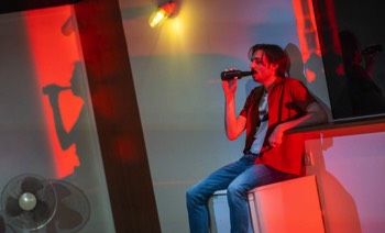 A Fight Against Production Image. Passerby (Eduardo Arcelus) sits on top of a half-height fridge drinking a beer. He is lit in bright red from beyond the picture. There is an outdoor wall lit illuminated to his right.