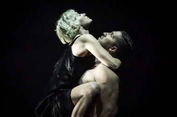  Julie (Heledd Gwynn) and John (Tim Pritchett) embracing in a tight spotlight. Julie is clinging on to John's upper body with her knees around his arms. The rest of the image is completely dark. John is topless with Julie in a black silk night gown. 