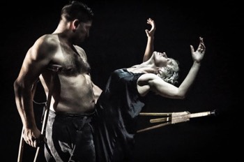 Julie (Heledd Gwynn) reclines backwards, her arms outstretched, over John's outstretched wooden crutch (Tim Pritchett). He supports his other side on another wooden crutch. He is topless wtih braces hanging to the sides of his brown period corded trousers. Julie wears a black silk night gown. They are lit tightly, the rest of the space is completely dark.