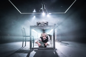  Julie (Heledd Gwynn) crawls under a long metal table. Bright lights shine directly behind her causing a spectacular glare of light in the haze behind her. There is a rectangle of LED lights hanging overhead and a large bell suspended in the centre. 