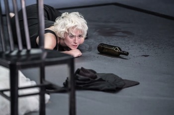  Julie (Heledd Gwynn) laying on the grey concrete like floor. There is an empty wine bottle beside her and splashes of wine all over the floor. In the foreground is a metal black chair and the floor has some strewn clothes across it. Julie is wearing a black silk night gown and has bright white permed hair. The stage is brightly lit in a cool white. 