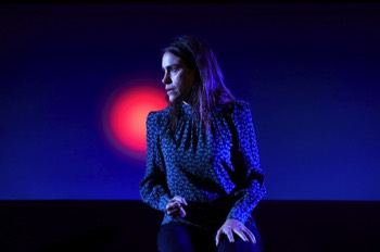 She (Emily Bruni) sits with her hands on her lap. She is looking to her right	 looking intently. There's a large screen behind her lit in dark blue from below, with a bright red dot on it over her right shoulder. She wears a blue floral dotted blouse and blue jeans.