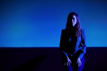  She (Emily Bruni) sits with her hands on her lap. She is looking blankly straight forward. She is only lit from her left in a cool blue colour, her right side is dark. There's a large screen behind her lit in light blue from below. She wears a blue floral spotted blouse and blue jeans. 