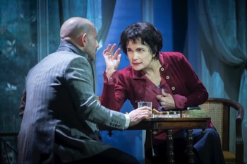Ava: The Secret Conversations production photo by Marc Brenner. Ava Gardner (Elizabeth McGovern) leans across a chess table to Peter Evans (Anatol Yusef). His back is to the camera, with an empty whiskey tumbler in his hand. There's an ashtray filled with cigarette butts on the table, a cigarette case and lighter. Ava studies Peter, her right elbow on the table and hand raised towards her head. Her other arm is clutched to her chest as a result of her stroke.