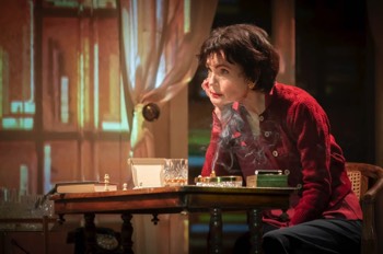 Ava: The Secret Conversations production photo by Marc Brenner. Ava Gardner (Elizabeth McGovern) sat at a chess table with her right elbow on the table, resting her chin on her hand. A cigarette smokes from the ashtray beside her. She looks intently beyond the image but also unamused. There's a partially drunk tumbler of whiskey on the table, along with a cigarette case, chess pieces and a book. The walls are projected with images of books. Ava wears a red cardigan over a red blouse.