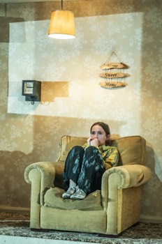 Wendy (Annabella Jennings) sits curled up on a worn out cream armchair in her tiny flat. An electricity meter is on the wall behind her and a cheap lamp shade hangs over head. Cold daylight streams in from the side.