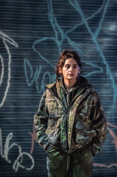 Sylvie (Ema Cavolli) stands in thick camouflage jacket against a graffiti covered roller shutter.