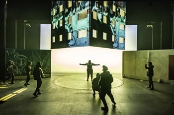 A wide shot of the stage, with Dennis (Tyreke Leslie) standing in the centre silhouetted by a bright yellow cyclorama. A skeleton of a tower block hangs overhead with its walls covered with projected video. Ensemble cast members are gathered around the stage edges.