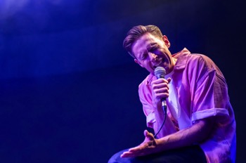 The Comedian (Samuel Barnett) sits on a stool in an open pink shirt with white tee under. He holds a mic to his mouth with his face screwed up in pain.