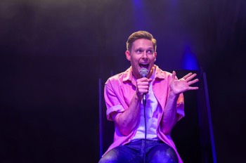 The Comedian (Samuel Barnett) sits on a stool in an open pink shirt with white tee under. He holds a mic to his wide smiling mouth and an open hand gesticulating to his side.