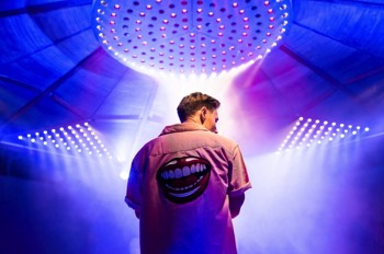 The Comedian (Samuel Barnett) stands with his back to the camera wearing an pink shirt with a large laughing mouth embroidered on the back. He looks down to his side. He is framed by a large arrays of LED lights, two triangles to his sides and a large circular array above him. The individual beams of the red, white and blue lights create patterns in the haze.