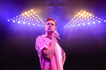The Comedian (Samuel Barnett) stands wearing an open pink shirt with white tee under. He holds a mic to his pursed mouth, his left hand on his hip. Large triangles of LED lights are behind him lighting up a cloud of haze.