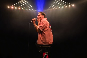 The Comedian (Samuel Barnett) ssitting on a stool away from camera, but turned to his left. He wears an open pink shirt with large smiling mouth embroidered on the back. He holds a mic to his mouth, his left hand clamping his jaw. Large triangles of LED lights are behind him lighting up a cloud of haze.