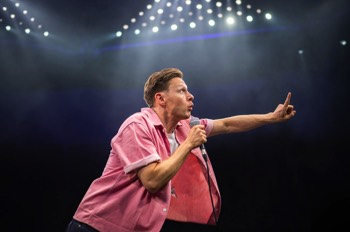 The Comedian (Samuel Barnett) stands wearing an open pink shirt with white tee under. He is lunging forward holding out a finger. Large triangles of cold white LED lights are behind him lighting up a cloud of haze.