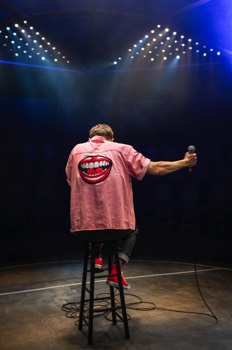 The Comedian (Samuel Barnett) sits on a stool with his back to the camera wearing an pink shirt with a large laughing mouth embroidered on the back. He's looking down with his shoulders hunched. He holds the microphone away out to his side. He is framed by large arrays of warm and cold LED lights lighting the haze in front of him.