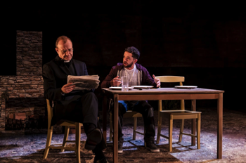 Clive (Tim Treloar) sits at the head of the table in his full vicar uniform reading a newspaper. Gary (Nathan McMullen) sits beside him looking incredulous, but Clive doesn't acknowledge him.