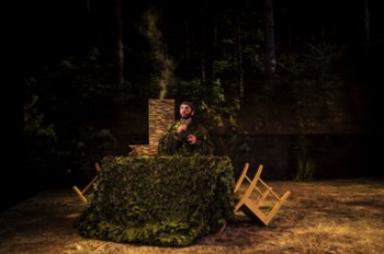 Reece (Alex Bhat) in full camouflage hiding in a hastily constructed lookout, with a projection of woodland behind him. Smoke rises out from his lookout as he looks upwards holding binoculars.