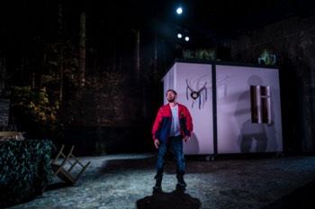 Gary (Nathan McMullen) cries out as he treads in dog poo, after bird poo has landed on his shoulder. The forest is dark with woodland projected behind him.