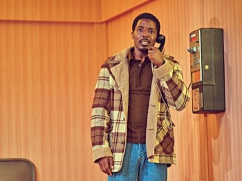 Jitney Production Image. Youngblood (Solomon Israel) talking on a payphone against a wood veneer panelled wall.
