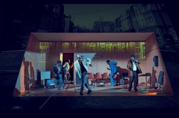 Jitney Production Image. The company dance to the radio in a darkened wood panelled office. The surrounding walls of the set are projected with a Pittsburg street which bleeds into the room.