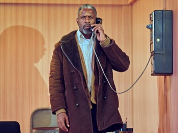 Jitney Production Image. Becker (Wil Johnson) stands on a payphone attached to the wood panelled office wall. He looks straight out towards the camera, looking concerned.