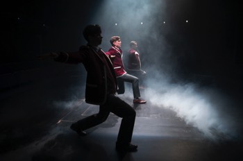 Three school boys in full uniform, surrounded by a low fog, are performing lunges in a line with their arms outstretched. They are lit in a single square cold white light from directly above.