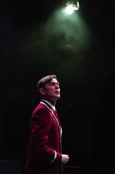 Harrison (Hubert Burton) stands in full school uniform, a red and gold tipped blazer. He is backlit in a single light, and is lit from out of frame by a cold white light. He looks off to the right, looking concerned.