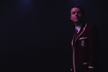 Lyons (Matthew Tennyson) stands in full school uniform looking outwards in the dark.