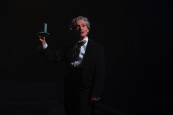 The Tour Guide (Eva Magyar) stands in a tailed black tuxedo jacket with dress shirt and large lace bowtie. She’s side lit in a single cold white light and holds a miniature monument in her right hand.