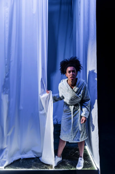 Vi (Jade Ogugua) peeks out from behind a large white drape wearing a white dressing gown and trainers.