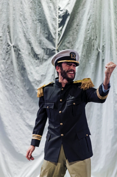 Leonard (Alex Bhat) wears a full navy uniform holding a fist up triumphantly.