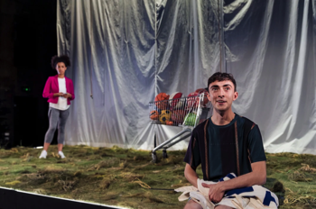 Laurie (Joe Barber) sits on the edge of grass stage looking out clutching a fabric banner. Behind him is a trolley full of basketballs and Vi looking on holding a mobile phone in her hands.