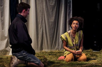 Laurie (Joe Barber) kneels on the grass stage beside Vi (Jade Ogugua) who sits on the grass in dirty clothes and a hi-vis jacket eating a cream bun.