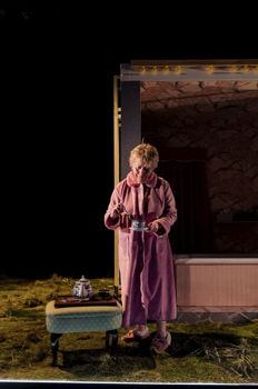 Margaret (Linda Broughton) stands in a pink dressing gown making a cup of tea over a tea tray on a green upholstered footstool set on the grass stage. Behind her is a dark enclosed bathroom.