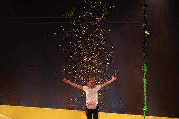  Purple Snowflakes and Titty Wanks Production Image. Saorise (Sarah Hanley) standing in the middle of the stage with an explosion of gold confetti firing over her from the floor behind. 