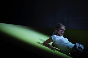  Purple Snowflakes and Titty Wanks Production Image. Saorise (Sarah Hanley) is laying on an inclined section of yellow carpeted set in a cold white spotlight. There's white powder smeared across one side of her face, and she is propping herself up on her elbows. 