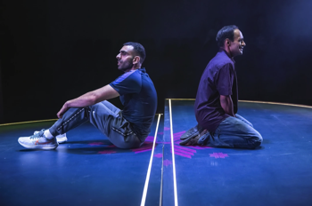 Bilal (Waleed Akhtar) sits on a the stage with his back to Zafar (Esh Alladi) who is kneeling. Two lines of light run along the floor between them.