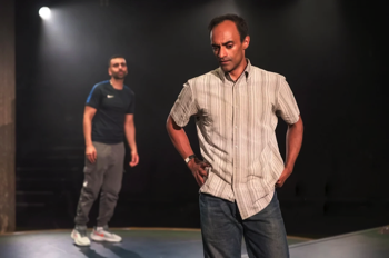 Zafar (Esh Alladi) stands in the foreground with his hands on his hips, looking at the floor. Bilal (Waleed Akhtar) stands in the background looking towards Zafar. They are lit in a neutral warm light.
