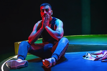 Bilal (Waleed Akhtar) sits on the edge of the circular stage with his arms on his knees holding his face. He's lit in cold light with a warm orange glow from the side.