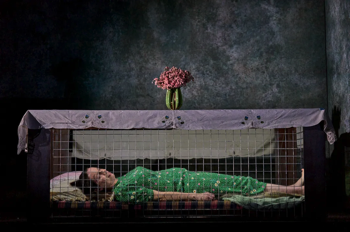 The woman (Kate O'Flynn) laying in the Morrison Shelter behind the mesh side. The top of the shelter is covered with a white table cloth and vase of pink flowers.