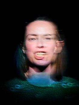 A blurred slow exposure headshot of the woman (Kate O'Flynn) speaking with only her face illuminated.