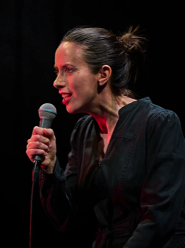 The woman (Kate O'Flynn) speaking into a microphone lit in red from behind.