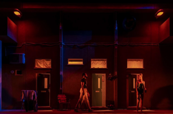 The whole stage during a transition is bathed in a hot orange light. Cast cross in front of the oppressive large concrete wall of the flats.