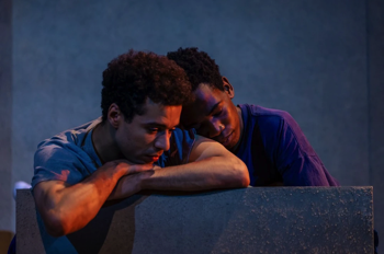 Jamie (Rilwan Abiola Owokoniran) rests his head on Ste's shoulder (Raphael Akuwudike) in his bed. They are dimly lit.