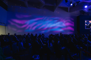 Lighting projections of blue and purple ripples across the congress centre's white walls, silhouetted audience sat below