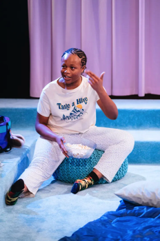 Funmi (Bukky Bakray) sits on a bean bag on a turquiose carpeted floor making a gun hand gesture to the side of her head.