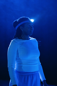 Elle (Shayde Sinclair) wearing an illuminated head torch, the rest of the space is dark, except for a low dark blue glow.