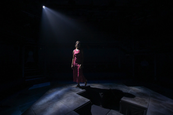 Bel (Ruby Crepin-Glyne) circling a cross shaped hole in the centre of the stage illuminated by a single light behind her.