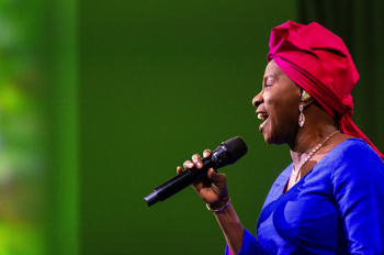 Angélique Kidjo singing wearing a red head dress and blue dress against a green projection. 